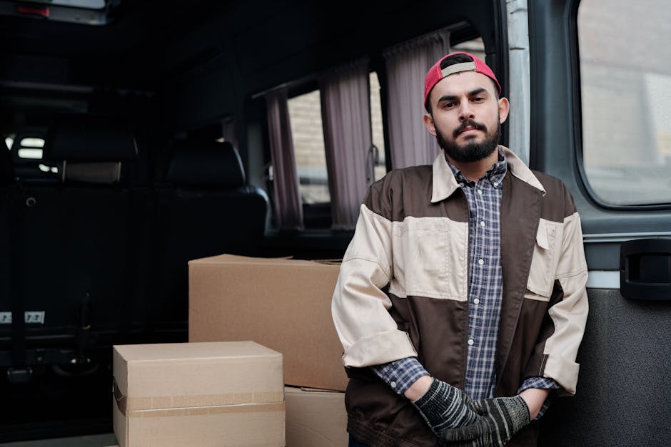 A man with a beard, wearing a red cap, a checkered shirt under a brown and beige jacket, and grey gloves, stands inside the rear of a van loaded with multiple cardboard boxes. The boxes are stacked neatly on the van’s floor, with some secured with packing tape, indicating a home relocation or moving process. The interior of the van features dark upholstery and curtains on side windows, with the outside environment visible through the window. This scene depicts the loading or preparatory phase of furniture transport and packing during a house removal service, highlighting the logistical aspects of delivery and transportation managed by Man and Van Lambeth, as part of their removals service category. Proper packing materials and careful handling are crucial for safe domestic removals, ensuring furniture and belongings are protected during the move.