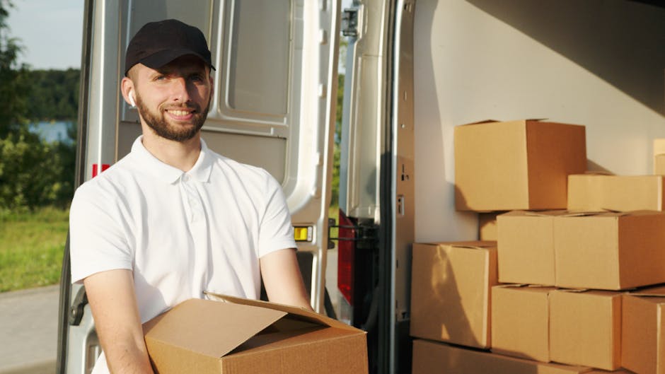A man wearing a white polo shirt and a black cap is standing beside an open removal van, holding a cardboard box. Inside the van, several other cardboard boxes are stacked neatly on the floor and against the side walls, ready for loading or unloading during a home relocation. The van is parked outdoors on a paved surface with a background of green trees and a partly cloudy sky, indicating a daytime moving process. The scene captures the loading or packing stage of furniture transport, with details including cardboard boxes, the vehicle interior, and the outdoor environment, reflecting professional removals services by Man and Van Lambeth as part of their house moving and packing logistics services.