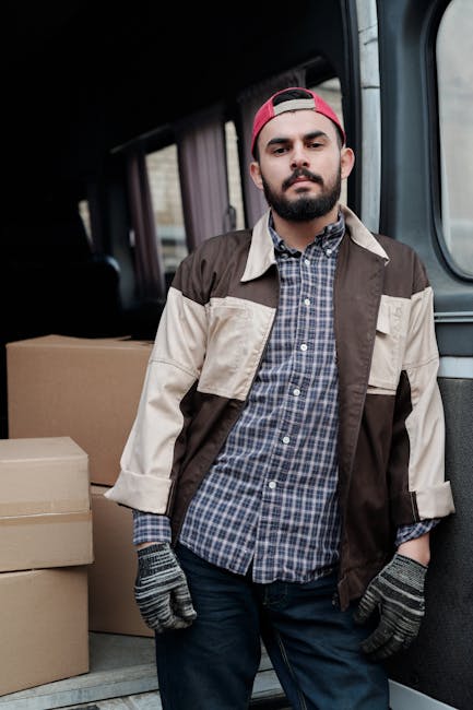 A man with a beard, wearing a red cap, a checkered shirt under a brown and beige jacket, and grey gloves, stands inside the rear of a van loaded with multiple cardboard boxes. The boxes are stacked neatly on the van’s floor, with some secured with packing tape, indicating a home relocation or moving process. The interior of the van features dark upholstery and curtains on side windows, with the outside environment visible through the window. This scene depicts the loading or preparatory phase of furniture transport and packing during a house removal service, highlighting the logistical aspects of delivery and transportation managed by Man and Van Lambeth, as part of their removals service category. Proper packing materials and careful handling are crucial for safe domestic removals, ensuring furniture and belongings are protected during the move.