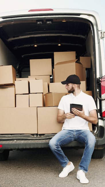 A man wearing a white t-shirt, blue jeans, and a black cap is sitting on the pavement next to an open-moving van, which is packed with numerous packed cardboard boxes of varying sizes, some sealed with packing tape. The boxes are arranged inside the van, with some positioned on top of others, ready for home relocation or furniture transport. The man is holding a tablet device, possibly managing the packing or moving schedule, while the interior of the van reveals a dark, spacious cargo area prepared for loading or unloading. The scene illustrates a loading process, with boxes inside the van and the man preparing for transport, in an outdoor setting with natural lighting. Man and Van Lambeth's removal services are implied through this context of packing and loading for a move near Waterloo station in Lambeth.