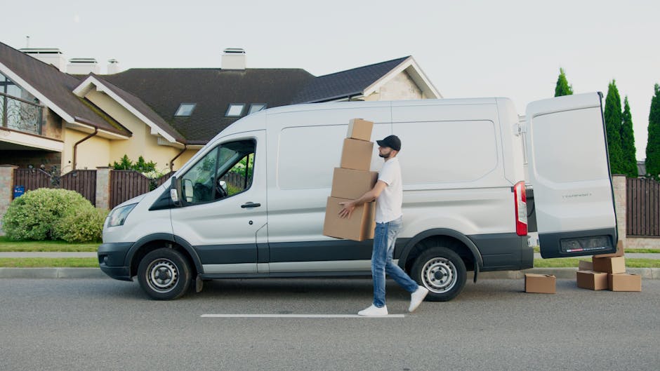 A man wearing a white t-shirt, jeans, and a black cap is unloading cardboard boxes from a white cargo van parked on a suburban street. He is carrying three stacked boxes towards the open rear door of the van, which is partially filled with other moving cartons. Several additional boxes are placed on the ground nearby, some resting on the pavement, indicating an ongoing home relocation process. The van, branded with black and gray accents, is positioned in front of a residential property with a house featuring a dark pitched roof, beige walls, a balcony, and a wooden fence in the background. Tall, green trees are also visible behind the house, and the scene is lit by daylight with clear weather. The image captures the loading process, involving furniture transport, packing, and moving logistics, as seen in the context of professional removals by Man and Van Lambeth, supporting house removals and relocation services near Waterloo station Lambeth.