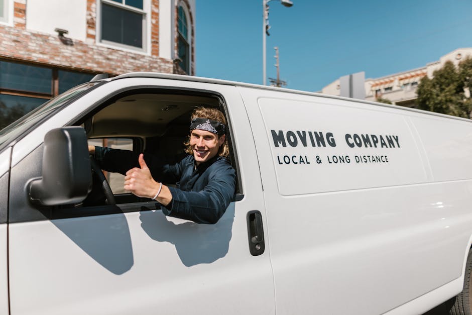 A man wearing a white t-shirt, blue jeans, and a black cap is sitting on the pavement next to an open-moving van, which is packed with numerous packed cardboard boxes of varying sizes, some sealed with packing tape. The boxes are arranged inside the van, with some positioned on top of others, ready for home relocation or furniture transport. The man is holding a tablet device, possibly managing the packing or moving schedule, while the interior of the van reveals a dark, spacious cargo area prepared for loading or unloading. The scene illustrates a loading process, with boxes inside the van and the man preparing for transport, in an outdoor setting with natural lighting. Man and Van Lambeth's removal services are implied through this context of packing and loading for a move near Waterloo station in Lambeth.