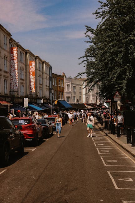 A narrow street in Brixton Market with multi-storey buildings on the left side, featuring colourful vertical banners and storefronts at ground level. The street is occupied by pedestrians walking along the pavement, some carrying shopping bags, while others are engaged in conversation or browsing the stores. Parked cars line the curb on the left, and to the right, there are designated parking bays separated by bollards and a pedestrian walkway. Large leafy trees extend over part of the street, providing shade. In the background, more buildings and market stalls are visible under a partly cloudy sky. This vibrant setting illustrates an active area suitable for local house removals and relocating belongings, with household items and furniture often transported through similar busy streets. Man and Van Lambeth offers comprehensive removals services, including packing, loading, and furniture transport, essential for home relocations in this busy Brixton neighbourhood.