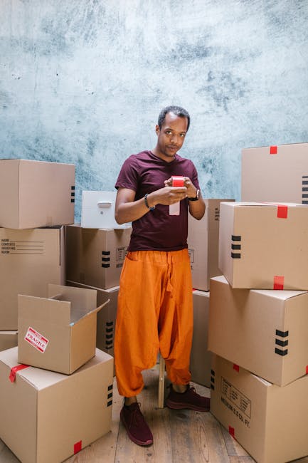 A man standing indoors among numerous cardboard moving boxes of various sizes, some sealed with red tape and others open revealing packing materials. He is looking down at his smartphone, wearing a maroon t-shirt and bright orange trousers, with a neutral expression. The background features a textured, light blue wall, and the floor is wooden. The scene depicts a home relocation or packing process, with the boxes arranged around him, potentially prepared for furniture transport and moving logistics. This setup exemplifies the packing and moving activities involved in house removals, aligning with services offered by Man and Van Lambeth.
