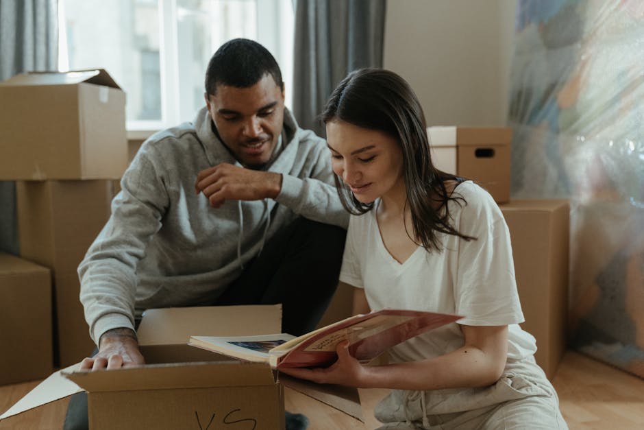 A woman with curly dark hair, wearing a beige sweater, is seated on a bed in a room with white walls, surrounded by packed cardboard boxes labelled with sizes such as 'Medium' and 'Large' and marked for office use; she is looking up and engaging with a person who is partially visible, holding a large box labelled 'BOOKS' and wearing a light-colored top and grey trousers, as they prepare for home relocation. The room contains a black metal shelving unit with wooden shelves, decorated with potted plants, and a closed window letting in natural light, indicating an organized packing and furniture transport process. The scene captures the loading and packing phase of a house move, with visible packing materials like tape and paper on the boxes, suggesting careful preparation for a professional removals service by [COMPANY_NAME] as part of their moving and packing logistics services.