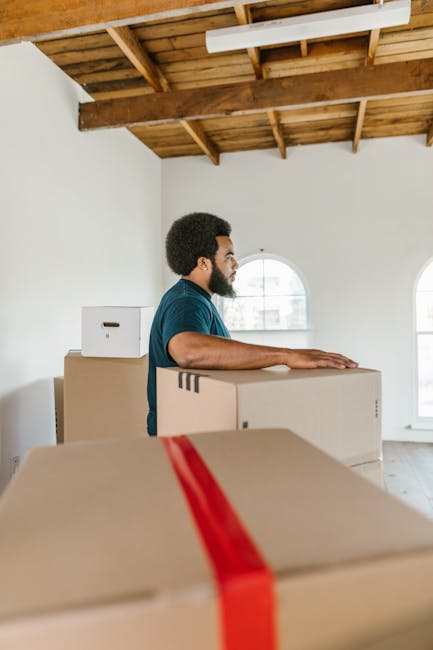 A man with dark curly hair and a beard, wearing a blue T-shirt, is standing in a spacious room with white walls and a wooden ceiling, engaged in a house relocation activity. He is holding a large card