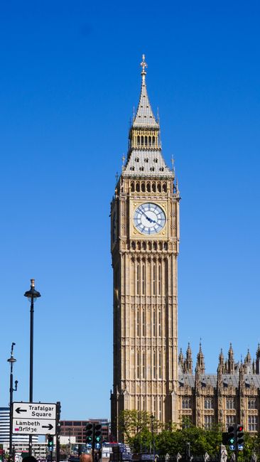 A tall clock tower known as Big Ben, situated on the Houses of Parliament in London, captured during daylight with a clear blue sky. In the foreground, a black street lamp and road signs indicating di