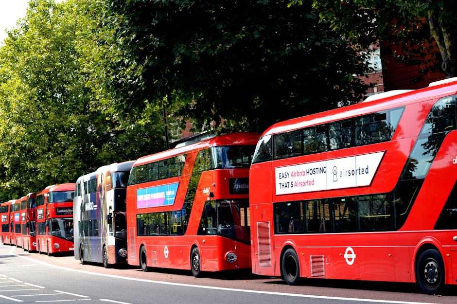 A row of red double-decker buses parked along a street with a background of green trees and part of a brick building. The buses are positioned side by side, with some visible advertisements on their sides, including one for Airsorted. The image captures the loading process of moving or relocating furniture and boxes, possibly outside a property, as part of home relocation services. The scene is well-lit, indicating daytime, and the surrounding environment suggests a London urban setting, relevant to house removals and transport logistics handled by Man and Van Lambeth.