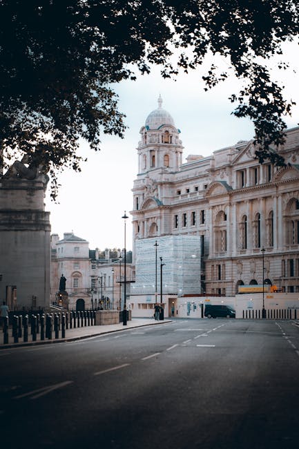 A street view of a historic building with classical architecture, featuring ornate stone facades, arched windows, and a prominent dome topped with a decorative spire, situated in the Lambeth area. The scene is framed by dark leafy tree branches at the top, casting partial shade over the empty road with minimal traffic and a few pedestrians. The building appears to be under some scaffolding or protective covering, indicating ongoing restoration work. Nearby are street lamps and bollards along the pavement, with a black van parked on the side. This setting is typical of an area involved in house removals and furniture transport activities, where careful planning and logistics are essential for moving belongings in and out of nearby properties. Man and Van Lambeth’s services often include facilitating such relocations, ensuring the safe loading and transport of furniture, boxes, and appliances during home relocation or packing and moving processes, especially around historic or central locations like this.