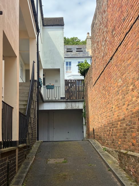 A narrow concrete driveway between a cream-colored residential building on the left and a red brick wall on the right, leading to a garage door beneath a small balcony with a black metal railing. The driveway has slight moss and dirt patches, with a few small weeds near the edges. On the balcony, there are potted plants, and a set of external stairs with black metal railings ascends on the left side of the cream building. In the background, upper floors of additional houses with grey roofs and white facades are visible, along with some greenery. This setting is part of a residential area suitable for house removals, where careful navigation and packing are essential due to the limited space between buildings, requiring managed furniture transport and precise loading processes. Man and Van Lambeth occasionally handles these logistical challenges as part of their removals service.