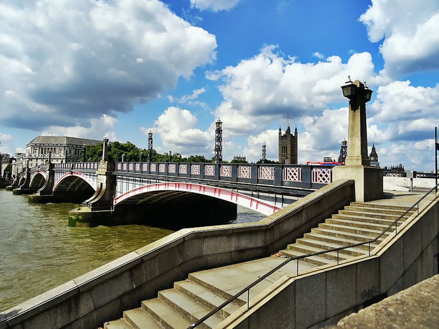 A view of Westminster Bridge over the River Thames in London under a partly cloudy sky, with the bridge featuring decorative lampposts and stone balustrades. In the background, the clock tower of Big Ben is visible, along with nearby historic buildings and various towers, indicating an iconic London cityscape. The foreground shows a stone staircase with metal handrails leading down to a pavement area adjacent to the riverbank, where a van from Man and Van Lambeth is parked for a house removal or furniture transport. The loading process involves the careful handling of packed moving boxes and furniture, some wrapped in plastic or blankets, with visible effort in the loading area to navigate the narrow stairs and limited parking space for efficient moving logistics. The scene captures the typical urban environment that presents parking and access challenges for removals, which Man and Van Lambeth reliably manages as part of their house relocation services. The overall environment suggests a professional setting for packing, transporting, and loading belongings for home relocation across London.