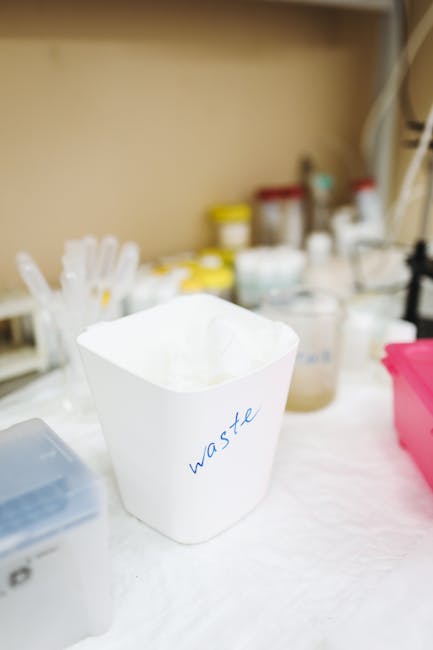 A white cardboard waste bin labeled 'waste' in blue handwriting, positioned on a white surface within an indoor setting, containing crumpled tissue paper or disposable material. In the background, there are various containers, bottles, and jars with yellow, white, and red caps, possibly used for cleaning, medical, or household supplies. The environment appears organised, with these items likely part of a packing or clearing process during house removals or home relocation. The scene is well-lit, with the focus mainly on the waste bin, supporting the context of sorting or disposing of packing materials as part of furniture transport and moving logistics, as handled by Man and Van Lambeth.
