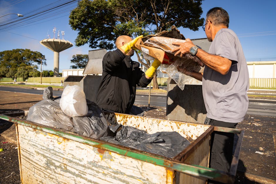 Two men are engaged in waste disposal outside a property, with one wearing black clothing and yellow gloves lifting a cardboard box filled with miscellaneous rubbish wrapped in plastic, while the other, dressed in a grey t-shirt, assists by holding the sides. They are positioned next to a large, rusty metal skip partially filled with black and grey plastic bags and other waste materials. The scene is set on a paved area adjacent to a street, under clear blue skies, with trees and a water tower visible in the background. An industrial-looking container with chipped paint is situated nearby, indicating an outdoor waste collection or removal process as part of a house relocation or moving service. This visual aligns with the logistics of packing, moving, and waste management for domestic or commercial removals, supported by Man and Van Lambeth's expertise in efficient furniture and rubbish disposal.